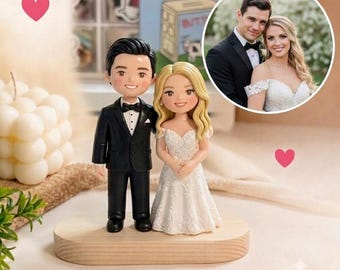 May include: A custom wedding cake topper featuring a miniature bride and groom figurine standing on a light wood oval base. The groom wears a black suit with a bow tie, and the bride wears a white lace gown. An inset photo shows the couple.