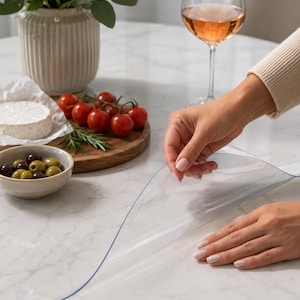 Può includere: Una protezione per tavolo in plastica trasparente e flessibile che viene posizionata su un piano bianco con motivo a marmo. Il materiale è liscio e sottile, progettato per coprire e proteggere la superficie rimanendo visibile sulla base.