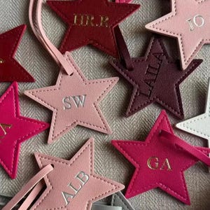 Może przedstawiać: Kolekcja zawieszek bagażowych w kształcie gwiazdek wykonanych ze sztucznej skóry w odcieniach różu, czerwieni i bieli. Każda zawieszka posiada pasujący pasek i jest wytłoczona spersonalizowanymi inicjałami lub imionami, takimi jak HR.R, JO, SW, LAILA i GA.