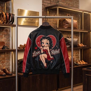 Puede incluir: Una chaqueta bomber negra con mangas rojas que presenta un gran gráfico en la espalda de un personaje de dibujos animados lanzando un beso dentro de un corazón rojo. El texto "Love Rebel" está impreso en letra cursiva bajo el corazón.