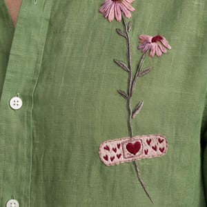 May include: A sage green button-down shirt featuring an embroidered design of two pink flowers on a stem, with a light pink adhesive bandage patch over the stem that includes a dark red heart and small red heart patterns.