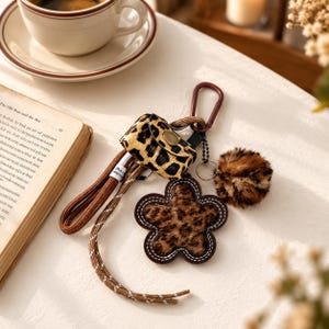 May include: A leopard print protective case for wireless earbuds featuring a brown carabiner, a matching leopard print faux fur flower charm, a fluffy leopard print pom-pom, and a brown braided lanyard cord with white stitching details.