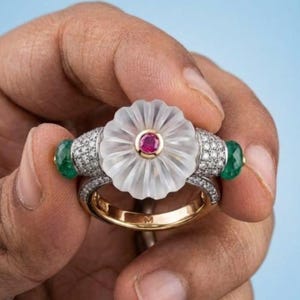 Può includere: Un anello in oro con un fiore centrale in quarzo trasparente intagliato e una piccola pietra preziosa rosa rotonda al centro. La fascia è decorata con piccoli diamanti bianchi e due perle di smeraldo verde rotonde ai lati della montatura.