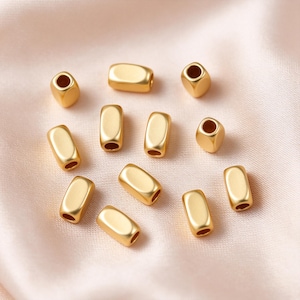 May include: A collection of twelve small, matte gold-toned beads with a smooth, rounded rectangular shape. Each bead features a central hole running through its length, showcasing a consistent metallic finish and uniform design across all pieces.