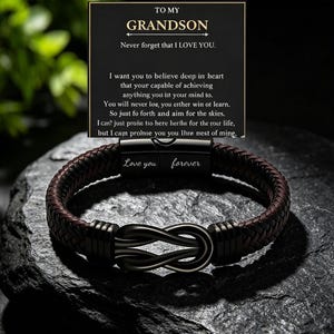 May include: A dark brown braided leather bracelet with a black metal infinity symbol centerpiece and a black clasp engraved with the words Love you forever. It is displayed with a black card featuring the text TO MY GRANDSON and an inspirational message.