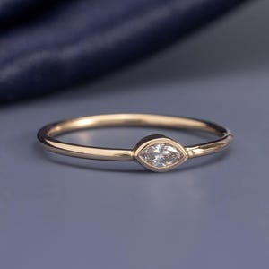 Könnte beinhalten: Ein dünner Goldring mit einem einzelnen, klar geschliffenen Edelstein im Marquise-Schliff, der horizontal in einer glatten Fassung sitzt. Der Ring hat eine polierte metallische Oberfläche und ein schlichtes, schmales Design.
