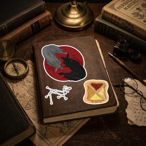 May include: Three stickers on a brown book cover: a circular sticker with a grey wolf and a black wolf against a red background, a white constellation sticker, and a square sticker of a toast slice with the text The Moody Toast.