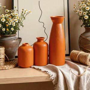 Peut inclure: Trois vases en céramique orange mat de hauteurs et de formes variées disposés en rangée. Les vases ont des surfaces lisses et des ouvertures étroites. Ils sont placés sur une surface en bois avec un tissu beige et une bobine de ficelle.