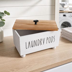 Può includere: Una scatola rettangolare in legno bianco con la scritta LAUNDRY PODS stampata in nero. La scatola presenta un coperchio in legno naturale con una piccola maniglia a pomello nera e poggia su quattro piccoli piedini arrotondati.