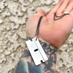 May include: A silver razor blade pendant necklace on a black chain. The pendant is hanging from a hand with tattoos.
