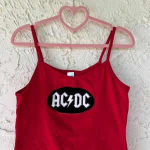 May include: A red spaghetti strap crop top featuring the band name "AC/DC" in white lettering inside a black oval with a lightning bolt. The top is hanging on a pink heart-shaped hanger.