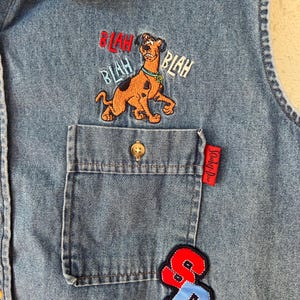 May include: A blue denim vest featuring an embroidered Scooby-Doo character above a chest pocket. The embroidery includes the text BLAH BLAH BLAH in red and blue. A red tag reads Scooby-Doo, and a red and blue SD patch is stitched below the pocket.