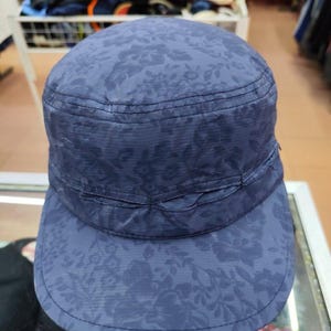 Puede incluir: Una gorra de estilo militar en color azul marino con un patrón floral tonal en toda la tela. La prenda presenta una parte superior plana, una corona estructurada y una visera corta y curva con costuras visibles en la banda y el borde.