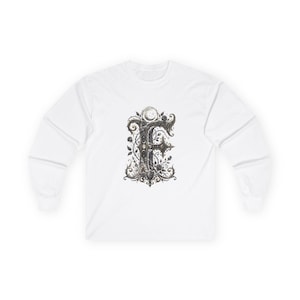 Op de afbeelding: Een wit shirt met lange mouwen en een grote, gedetailleerde, donkergekleurde letter F in het midden van de borst. De letter is versierd met gedetailleerde bloemranken, doornen en een kleine maanfasengrafiek bovenaan in grijstinten en zwart.