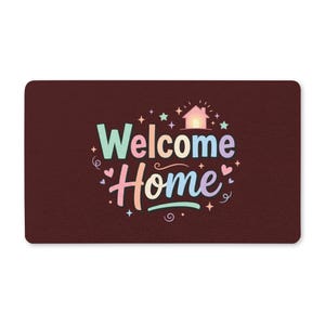 Op de afbeelding: Een rechthoekige donkerbruine deurmat met de tekst Welcome Home in een veelkleurig pastellettertype. Het ontwerp bevat een klein huisicoon, diverse sterren, hartjes en zwierige decoratieve lijnen rondom de centrale tekst.