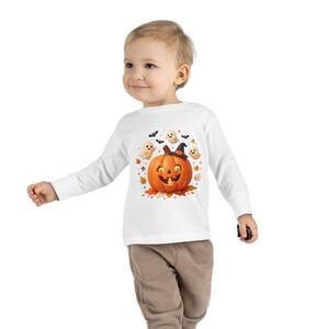 Op de afbeelding: Een wit shirt met lange mouwen voorzien van een centrale afbeelding van een lachende oranje pompoen met een zwarte heksenhoed, omringd door kleine witte spookjes, zwarte vleermuizen, herfstbladeren en maissnoepjes op een witte achtergrond.