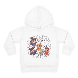 Op de afbeelding: Een witte hoodie met een centrale grafische print van zes stripfiguren in Halloween-kostuums, waaronder een heks, een pompoen, een geest, een vleermuis, een mummie en een duivel, omringd door kleine kleurrijke sterretjes en confetti.