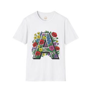 Op de afbeelding: Een wit T-shirt met korte mouwen en een grote, dikgedrukte letter A in het midden. De letter is gevuld met een gedetailleerde, kleurrijke illustratie van diverse bloemen, waaronder rode klaprozen, gele boterbloemen en paarse bloesems.