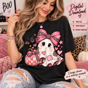 Op de afbeelding: Een zwart t-shirt met een afbeelding van een witte geest die een grote roze strik met luipaardprint draagt, zittend naast een roze en wit geblokte pompoen. Een kleine zwarte vleermuis en roze hartjes omringen de geest.