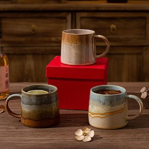 May include: Three ceramic mugs with reactive glaze finishes in shades of brown, beige, and blue. One mug sits atop a bright red square box, while two others are placed on a wooden surface. Each mug features a curved handle and a smooth rim.
