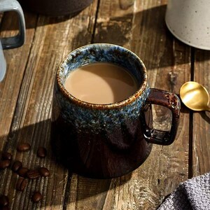 Puede incluir: Una taza de cerámica llena de una bebida de color marrón claro, con una base marrón oscuro que transiciona hacia un borde texturizado en azul y marrón. La taza tiene un asa resistente y un acabado brillante con un esmalte reactivo superior.