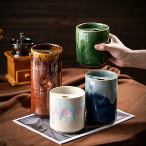 May include: Four cylindrical ceramic containers with a unique top shelf design. One is tall and rust-colored, one is green, one is blue and white, and one is cream with a pink drip glaze. Each features a small circular opening on the top ledge.