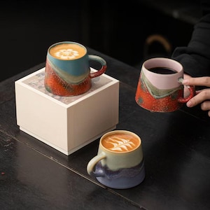 May include: Three ceramic mugs with textured, multi-coloured glaze finishes. One mug sits on a square box, one is held, and one rests on the surface. The mugs feature distinct colour combinations of blue, orange, pink, and purple with latte art.
