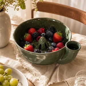 May include: A dark green ceramic berry bowl with a central drainage cone and a side handle, filled with fresh red strawberries and dark blue blackberries. The bowl has a glossy, speckled glaze finish and a wide, circular rim.