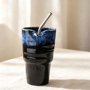 May include: A tall ceramic tumbler with a glossy finish, featuring a deep black base that transitions into a vibrant blue rim. The cup has a textured, rippled surface and a notched edge to hold a single, angled stainless steel drinking straw.
