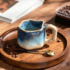 May include: A ceramic mug with a wavy, irregular rim and a gradient glaze transitioning from deep blue at the top to light beige at the base, featuring gold speckles. The handle is wrapped in natural jute twine, accompanied by a gold-toned spoon.