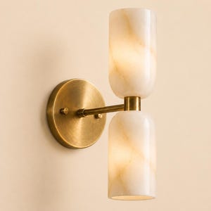 May include: A wall sconce featuring a circular brushed brass backplate and a horizontal arm supporting two vertically oriented, cylindrical white stone shades with subtle beige veining. The light fixture emits a soft glow from both stone cylinders.