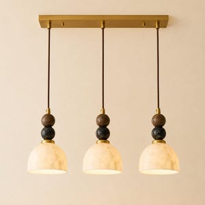 May include: A linear pendant light fixture featuring three dome-shaped white marble shades suspended from a rectangular brass ceiling plate. Each light includes a dark brown wooden sphere and a black marble sphere above the shade on a thin cord.