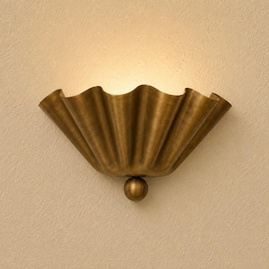 May include: A wall-mounted sconce light fixture featuring a wide, semi-circular shade with a pleated, fan-like design in a brushed brass finish. A small, spherical brass ball is attached to the bottom center of the fixture, which emits a warm glow.