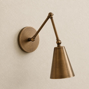 Puede incluir: Una lámpara de pared con brazo articulado y acabado en latón cepillado. El aplique cuenta con una placa circular para la pared, un brazo ajustable de dos piezas con articulaciones y una pantalla cónica orientada hacia abajo para dirigir la luz.