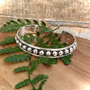 May include: A silver cuff bracelet with a row of small, round, silver beads. The bracelet is displayed on a wooden surface with green fern leaves. The bracelet has an open design.
