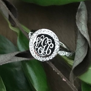 May include: A silver ring with a round, clear stone setting. The stone is surrounded by smaller clear stones. The ring has a monogram engraved in the center of the stone.