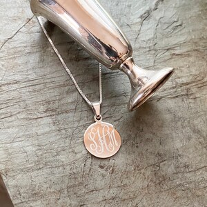 May include: A silver chain necklace with a round pendant featuring an engraved monogram. The pendant is hanging from a silver cup.