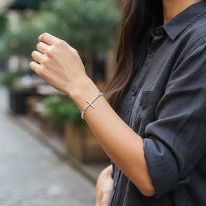Puede incluir: Pulsera de plata con un colgante de cruz. La pulsera est&aacute; hecha de peque&ntilde;as cuentas redondas. La cruz tiene un dise&ntilde;o sencillo. La persona lleva una camisa gris oscuro abotonada. Una joya elegante.