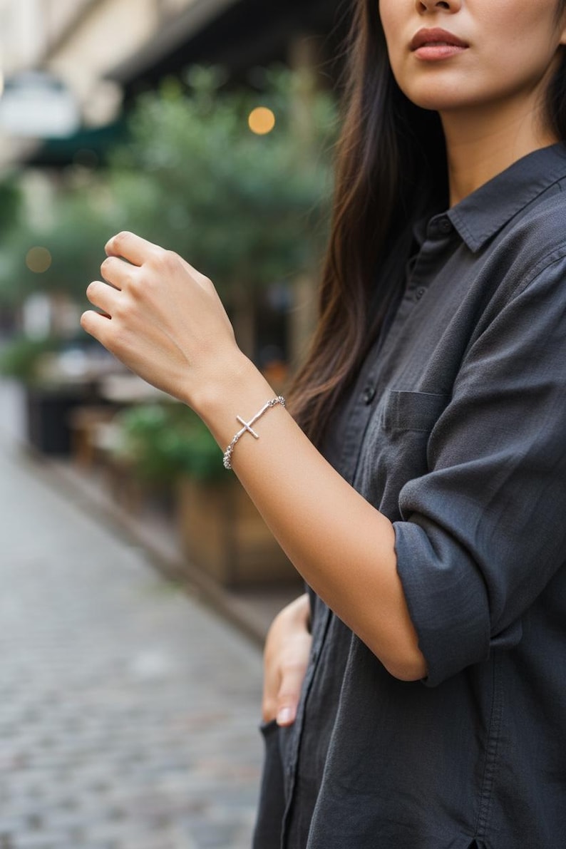 Puede incluir: Pulsera de plata con un colgante de cruz. La pulsera est&aacute; hecha de peque&ntilde;as cuentas redondas. La cruz tiene un dise&ntilde;o sencillo. La persona lleva una camisa gris oscuro abotonada. Una joya elegante.
