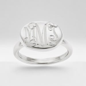 May include: A silver ring with a round face featuring an engraved monogram. The ring has a polished finish and a simple band. The monogram appears to be three intertwined letters.
