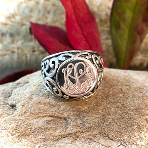May include: A silver ring with an ornate filigree design. The ring features a circular center with engraved initials. The ring is set against a natural stone background, with red and green foliage in the background.