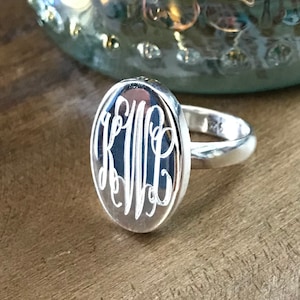 May include: A silver oval ring with a monogram engraved on the top. The ring is on a wooden surface.