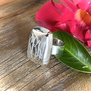 May include: A silver ring with a rectangular, engraved top. The ring is on a wooden surface with a pink flower petal and a green leaf.