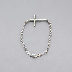 Puede incluir: Una pulsera plateada con un colgante en forma de cruz. La pulsera presenta una cadena de peque&ntilde;os eslabones redondos y un cierre. El dije de cruz tiene un dise&ntilde;o texturizado y org&aacute;nico.