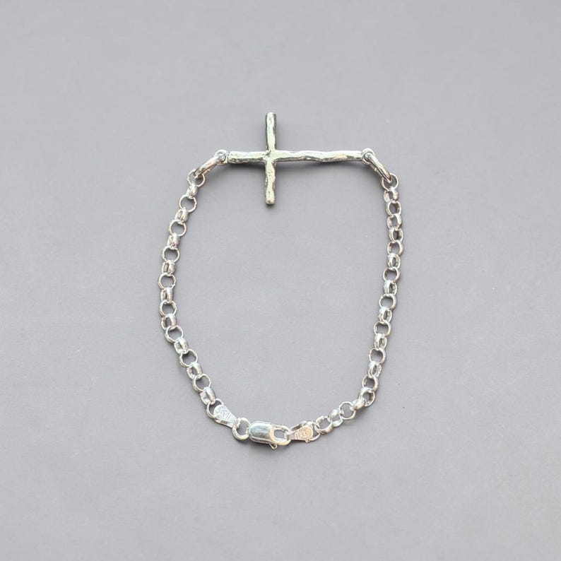 Puede incluir: Una pulsera plateada con un colgante en forma de cruz. La pulsera presenta una cadena de peque&ntilde;os eslabones redondos y un cierre. El dije de cruz tiene un dise&ntilde;o texturizado y org&aacute;nico.