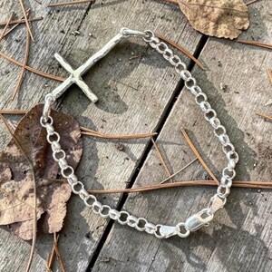 Puede incluir: Una pulsera de plata con un colgante de cruz. La pulsera presenta un dise&ntilde;o de eslabones de cadena y una cruz texturizada. El cierre es visible. La pulsera se muestra sobre una superficie de madera con hojas y agujas de pino.