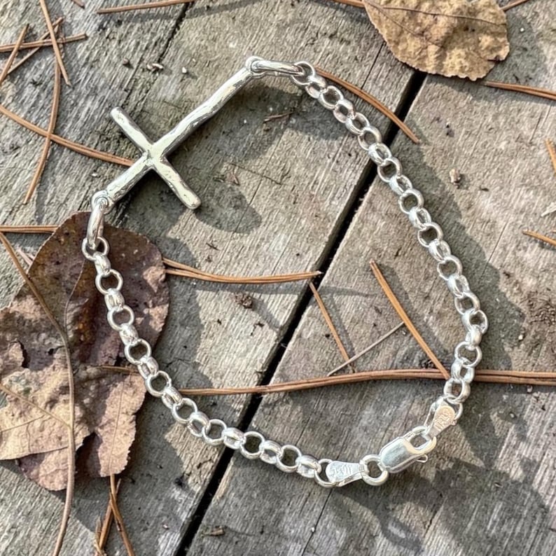 Puede incluir: Una pulsera de plata con un colgante de cruz. La pulsera presenta un dise&ntilde;o de eslabones de cadena y una cruz texturizada. El cierre es visible. La pulsera se muestra sobre una superficie de madera con hojas y agujas de pino.