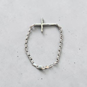 Puede incluir: Una pulsera plateada con un colgante de cruz. La cruz tiene un dise&ntilde;o texturizado y org&aacute;nico. La pulsera es de tipo cadena con cierre de mosquet&oacute;n. La joya se exhibe sobre una superficie gris.