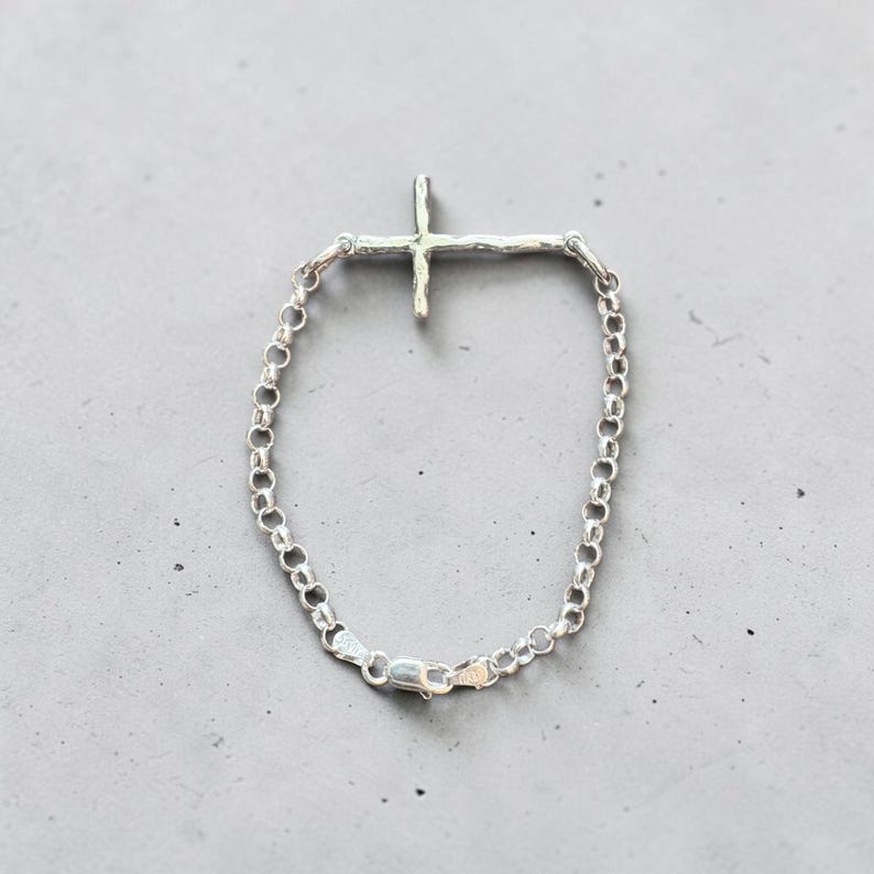 Puede incluir: Una pulsera plateada con un colgante de cruz. La cruz tiene un dise&ntilde;o texturizado y org&aacute;nico. La pulsera es de tipo cadena con cierre de mosquet&oacute;n. La joya se exhibe sobre una superficie gris.