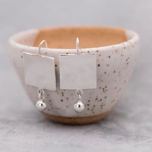 May include: A pair of silver earrings with square-shaped pendants and small round beads hanging from them. The earrings are displayed in a white ceramic bowl.